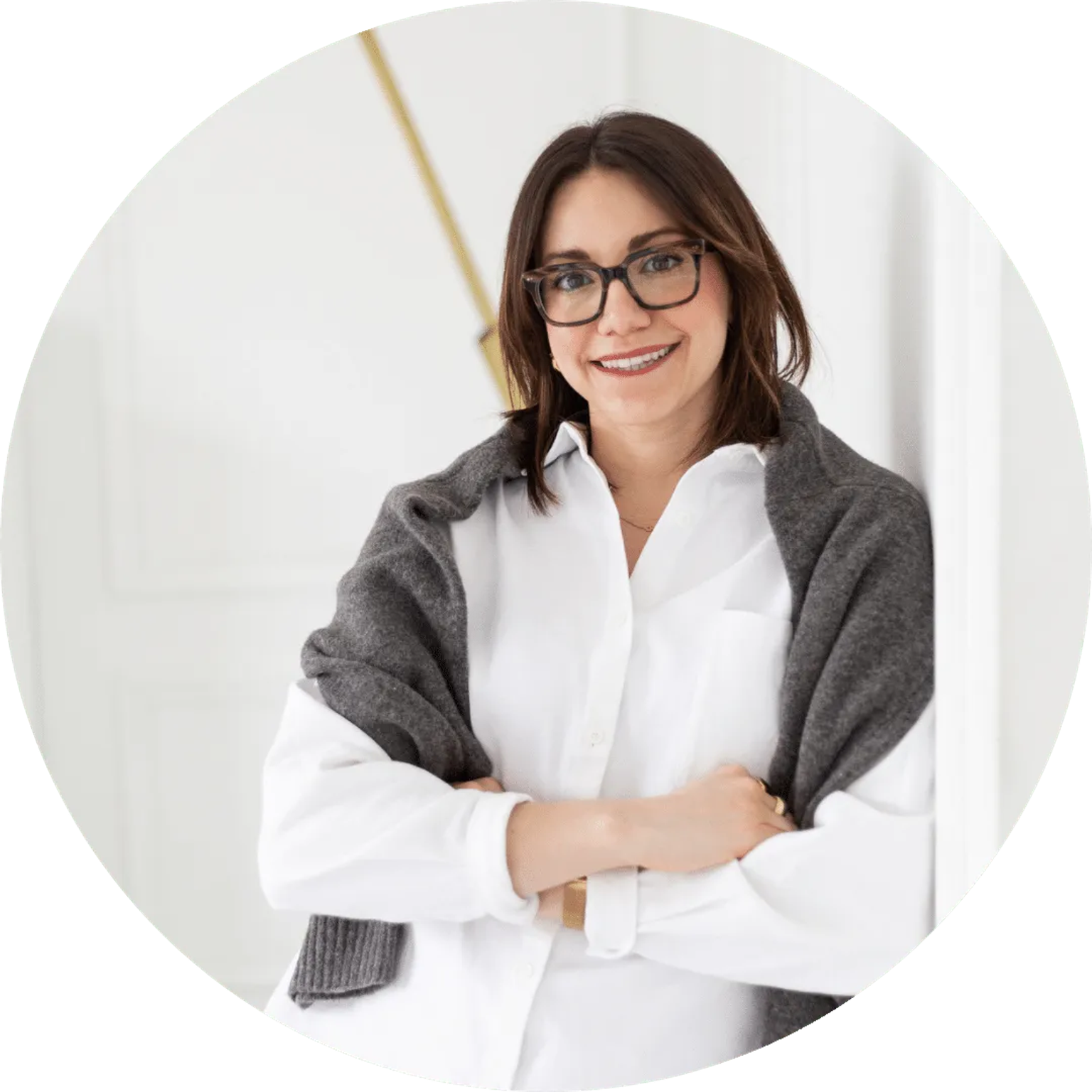 Smiling woman with glasses in a white shirt, wearing a gray cardigan, arms crossed against a light background.