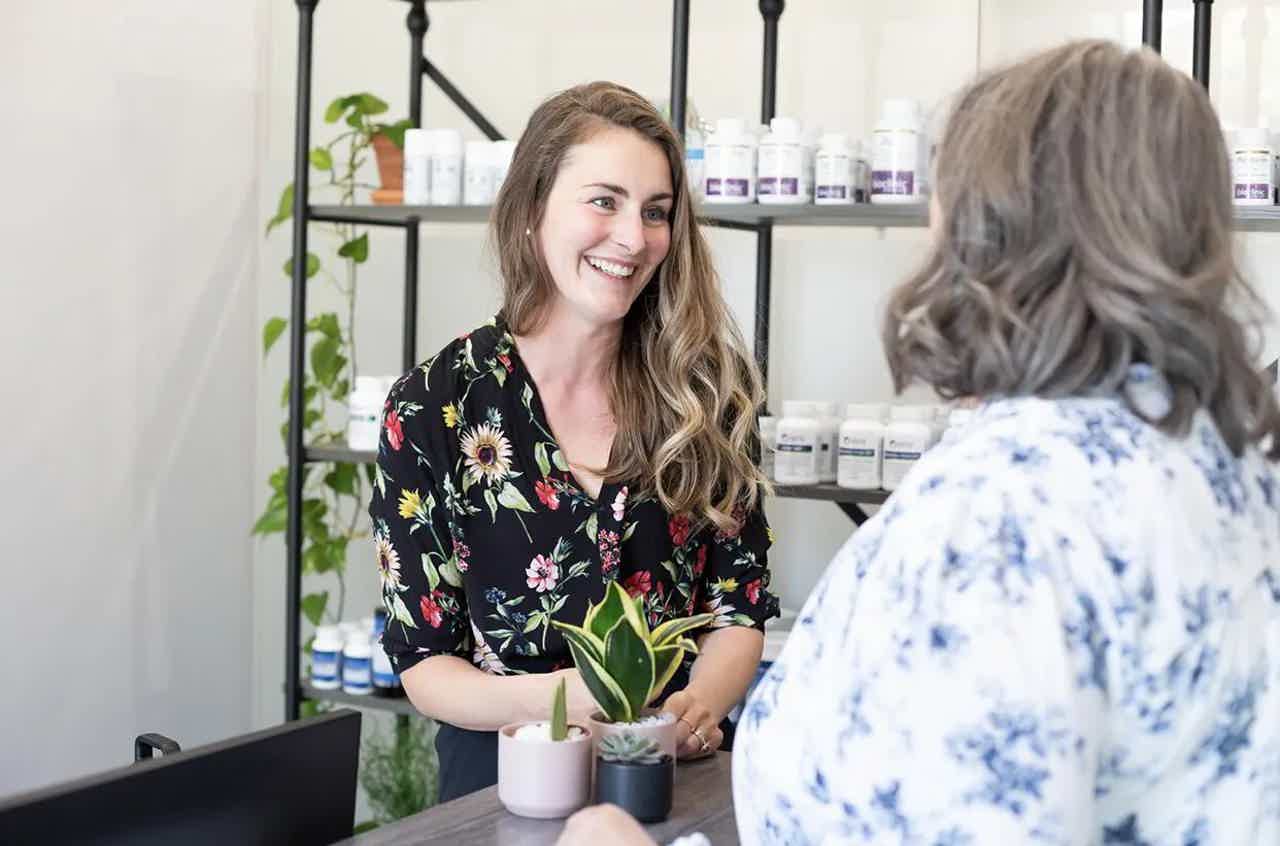 Woman with long hair smiles while talking with a seated customer in a beauty shop, surrounded by skincare bottles.