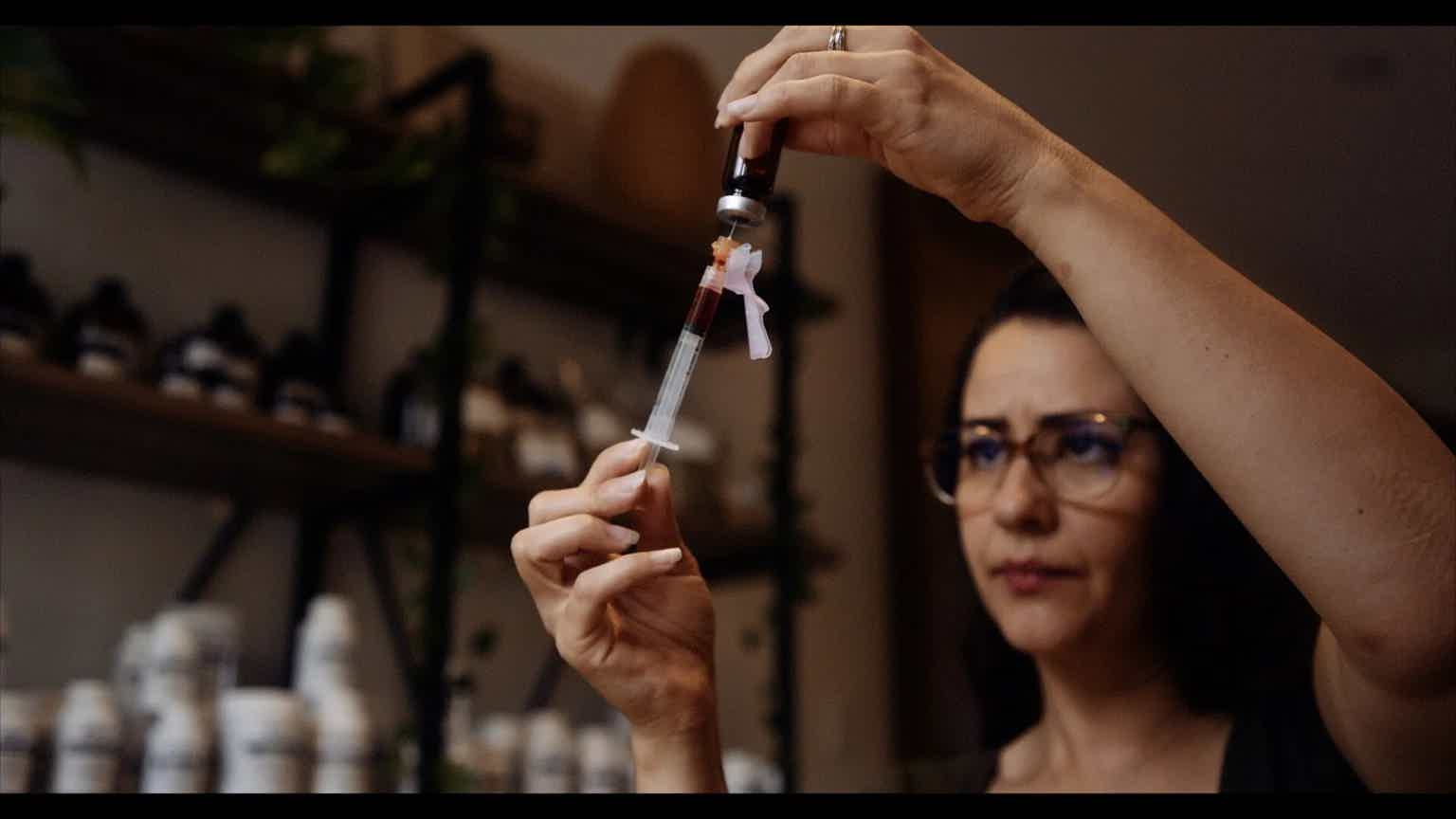 Woman raises a syringe over a seated person in a blurred medical setting; clinician holds needle and tubing.