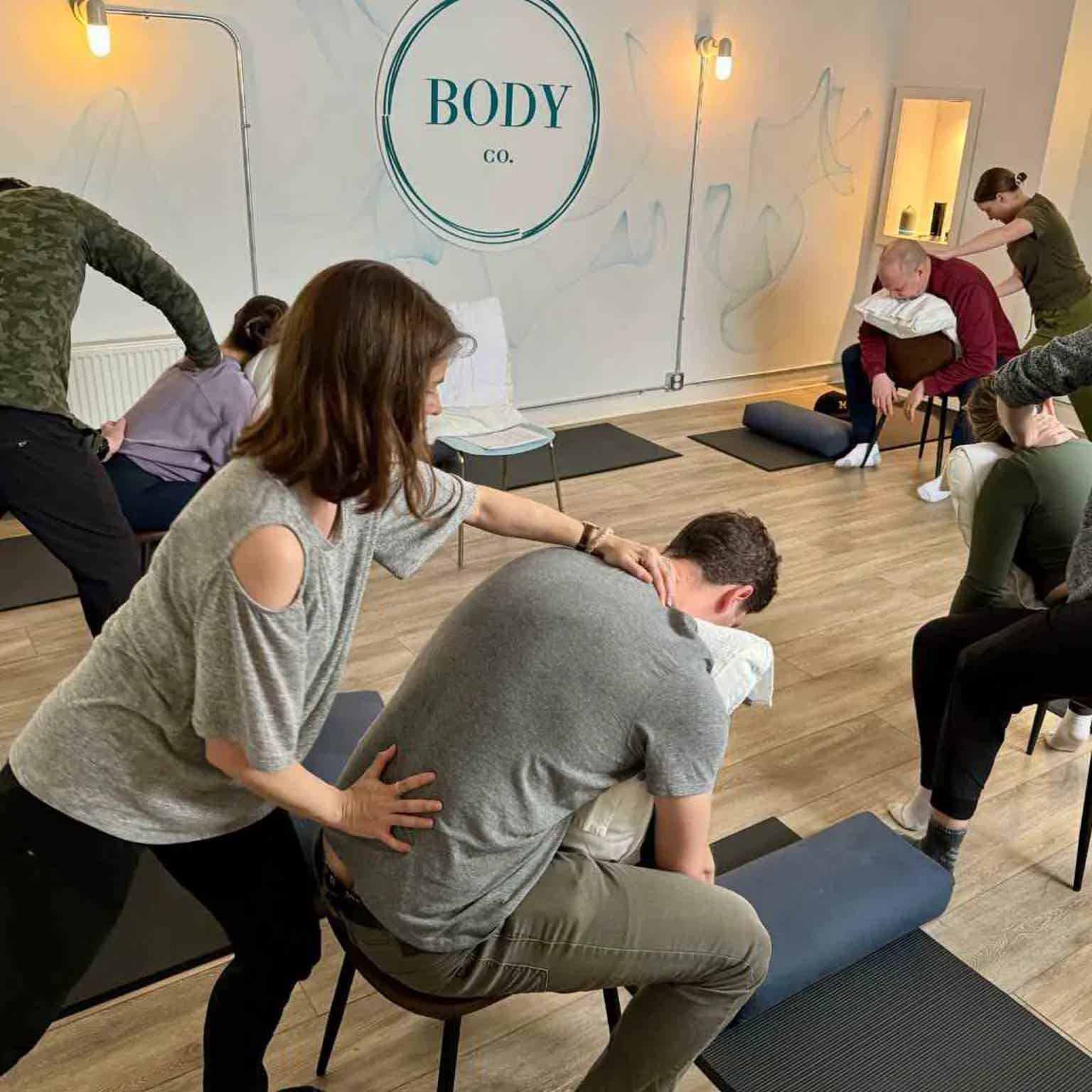 BodyCo classroom with therapist assisting seated participant on a mat while others stretch in the background.