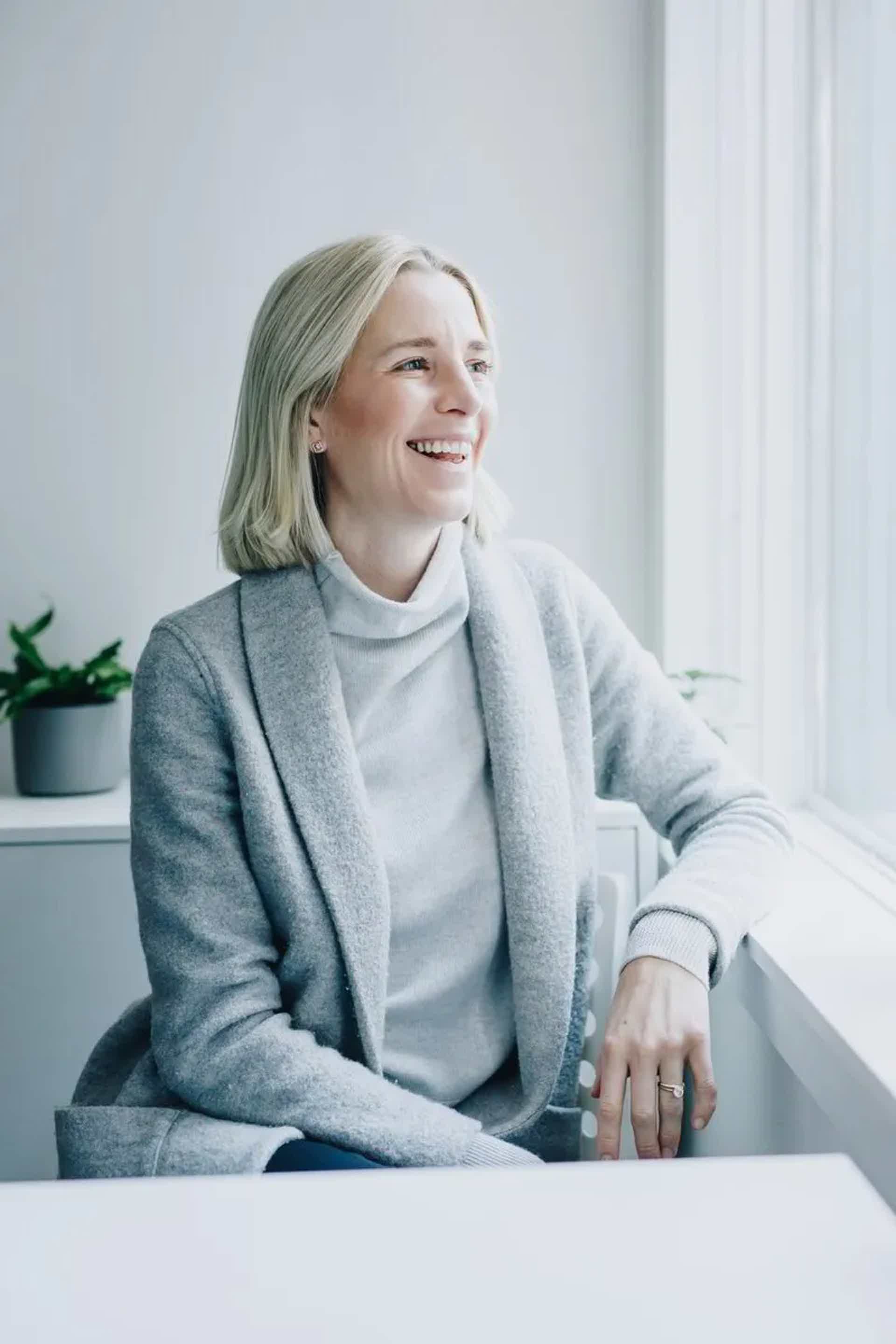 Smiling woman with gray hair sits by window, wearing a light gray coat and turtleneck, gazing outward.