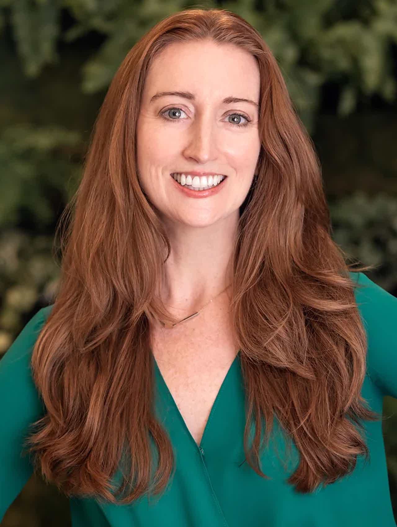 Smiling woman with long wavy red hair wearing a green top, outdoors with greenery in the background.
