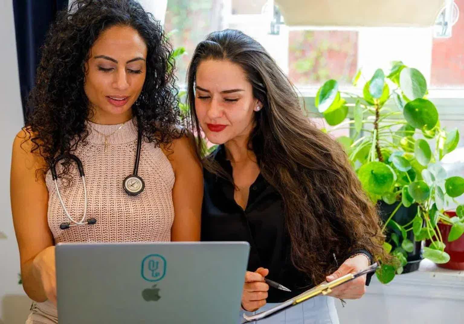 Two women collaborating at a desk, reviewing a laptop while one writes on paper near potted plants.