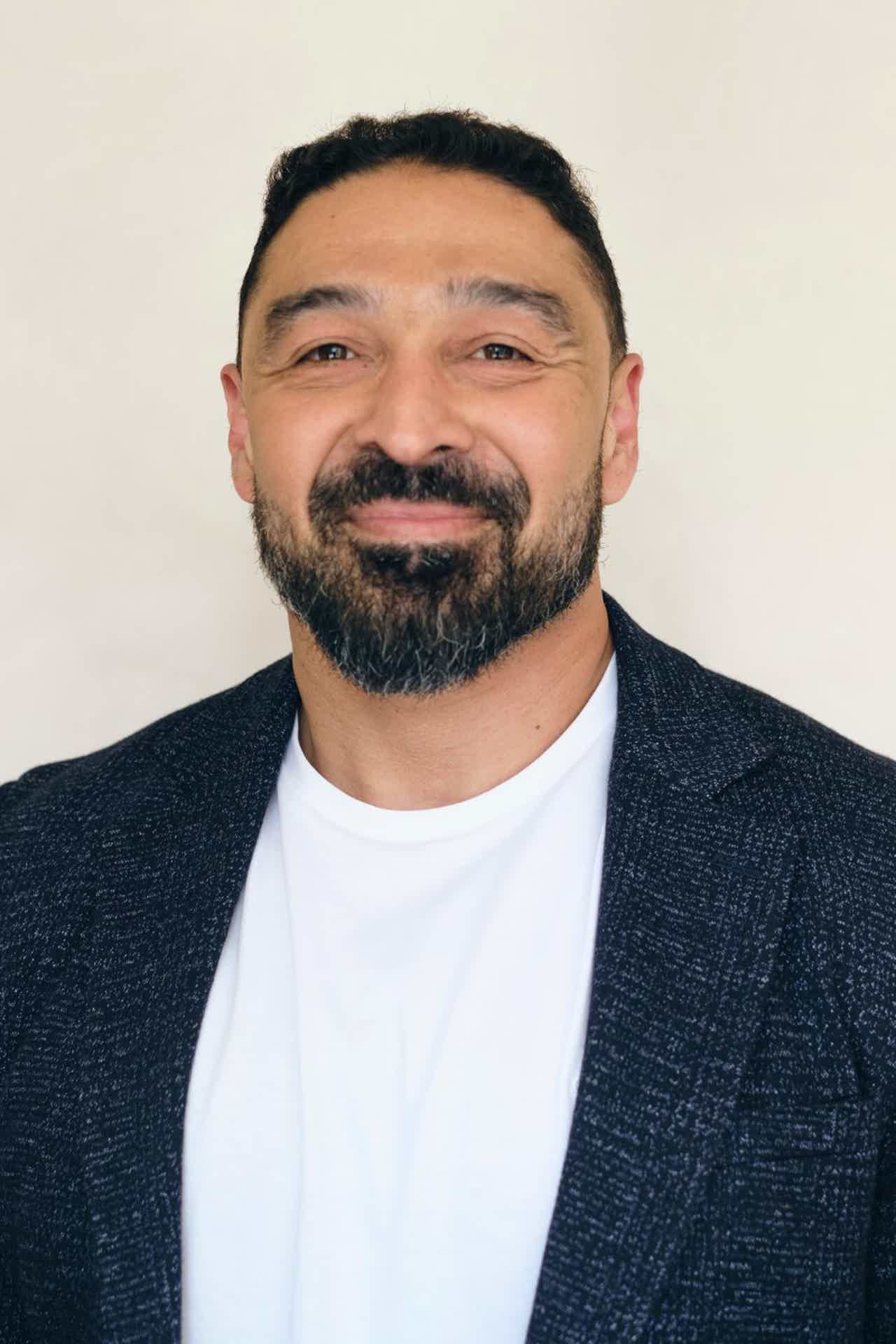 Smiling man with dark hair and full beard wearing a dark blazer and white shirt against a light background.