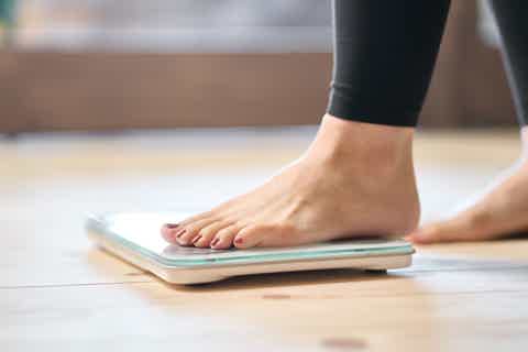 Bare feet stepping on a bathroom scale on a wooden floor, near a bed with soft blur background.