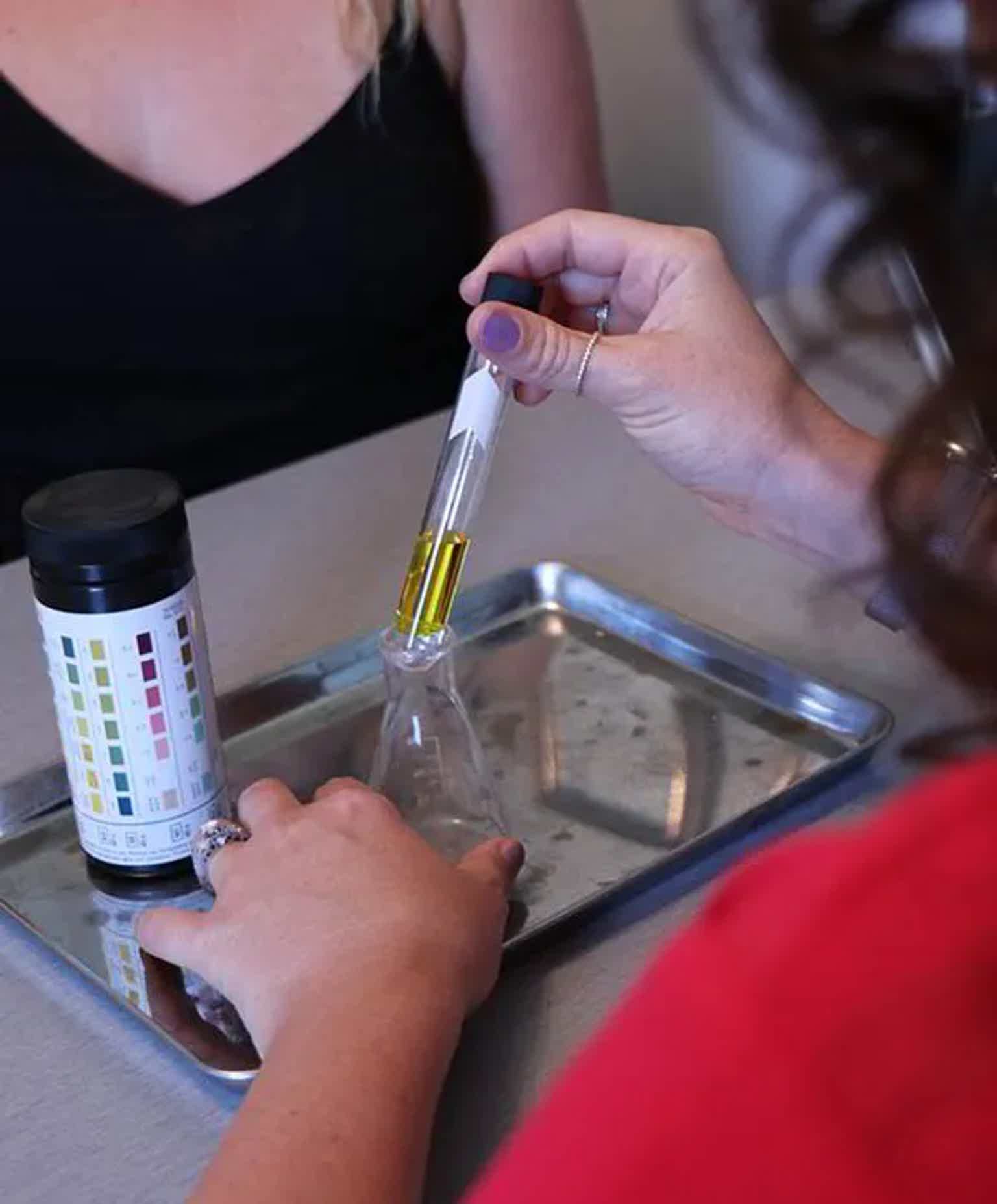 Two hands conducting a liquid test: a dropper transfers yellow liquid into a conical flask on a metal tray; color chart and c