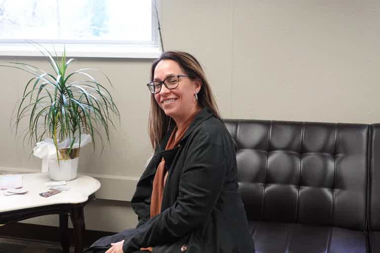 Smiling woman with glasses sits on a black tufted sofa in a room with a potted plant and small table.