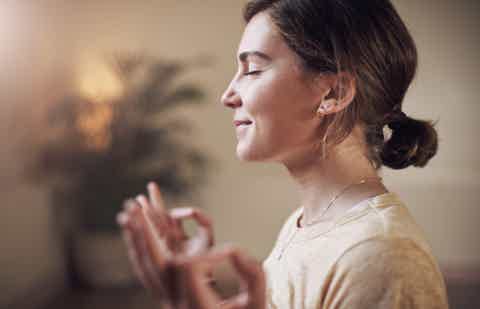 Side profile of a woman with hands raised in meditation, warm light, soft blurred background.