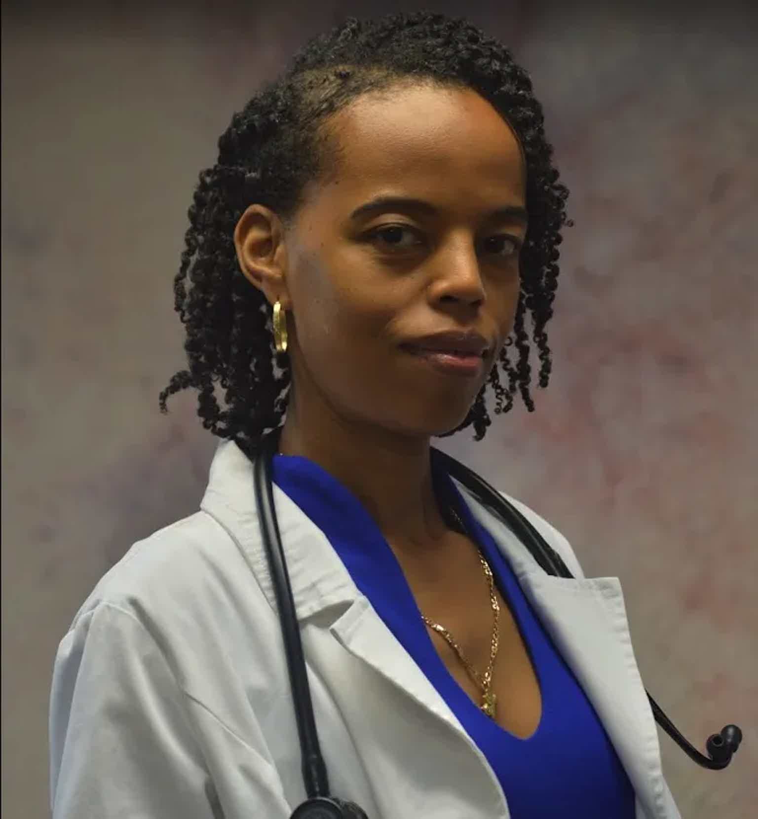 Caucasian woman with short twists in a white lab coat and stethoscope, blue top, gold jewelry, confident expression.