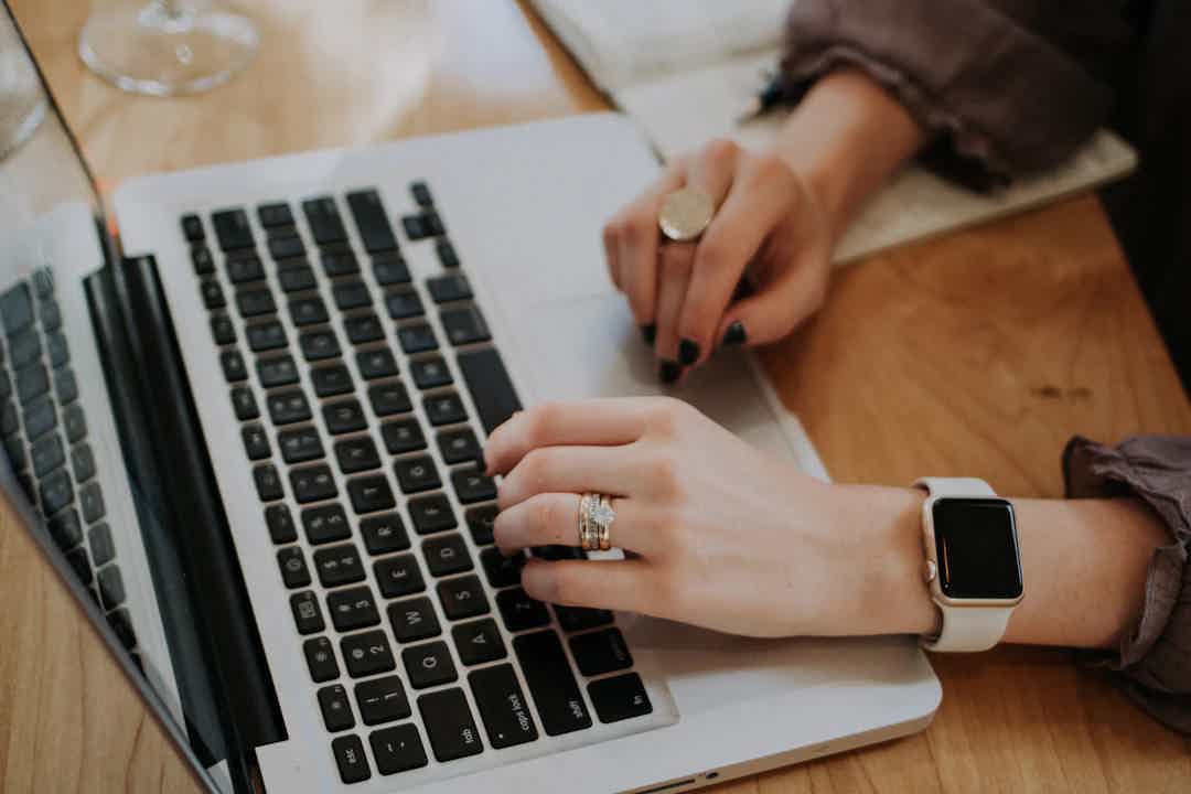 Hands typing on laptop keyboard with smartwatch nearby, desk setting and another hand holding a stylus