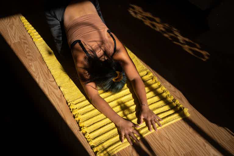 Person on a yellow rug doing forward fold stretch, arms extended toward floor, wooden floor and shadows around.