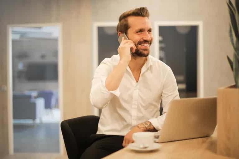 Man in white shirt smiles while talking on a smartphone at a desk with a laptop and coffee cup.