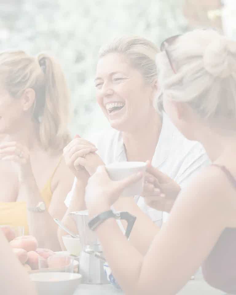 Friends laughing and chatting around a table with coffee and fruit, bright outdoor setting.