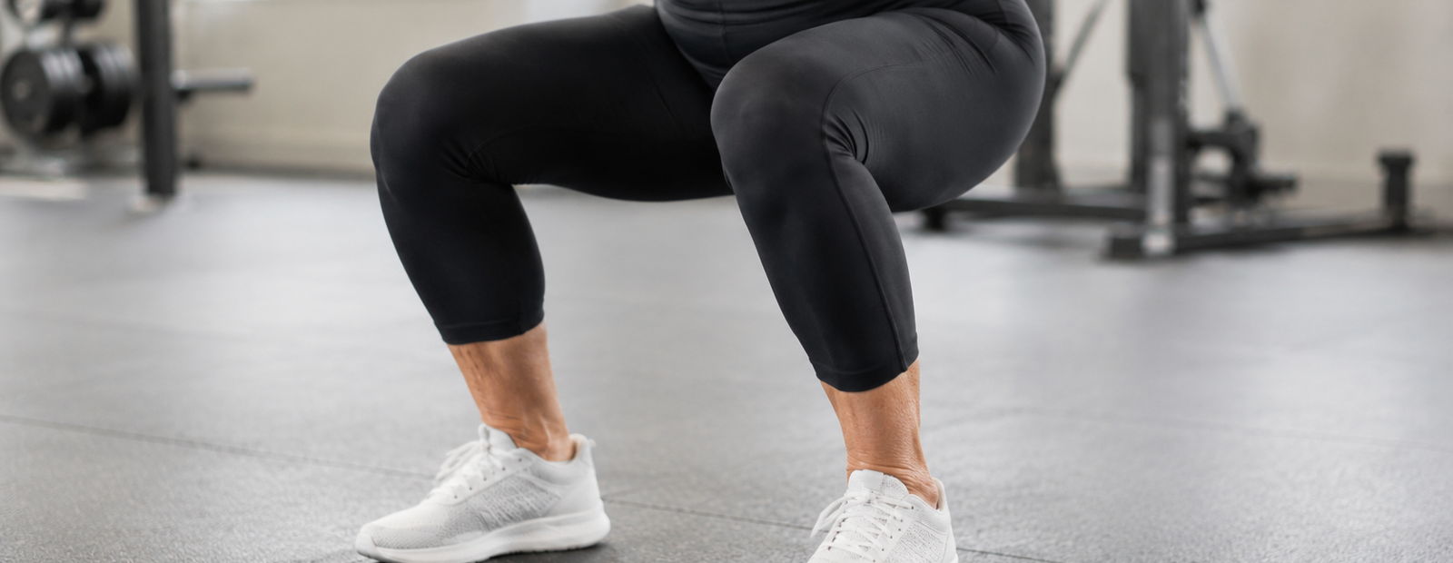 Person in black leggings and white sneakers doing an exercise in a gym, knees bent and body lowered.