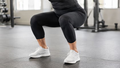 Person in black leggings and white sneakers doing an exercise in a gym, knees bent and body lowered.