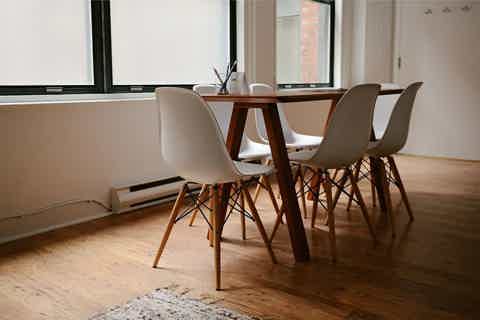 Minimalist dining area with wooden table, four beige shell chairs, and large windows letting in daylight.