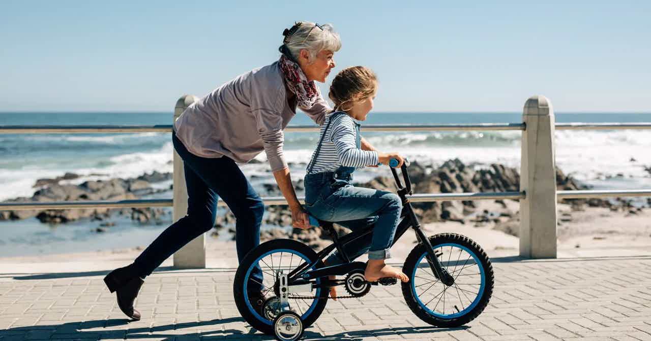 Older person helping a child ride a small bicycle on a seaside promenade with ocean waves behind them.