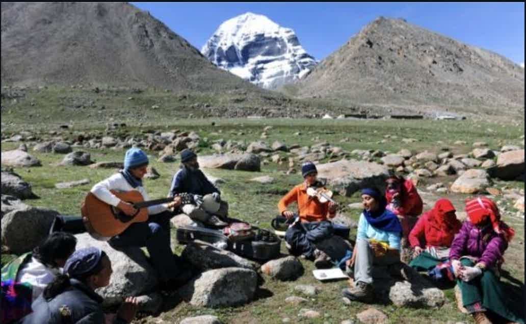 Group of musicians in a mountain valley with sheep herders, camp, and snowcapped peak in background.