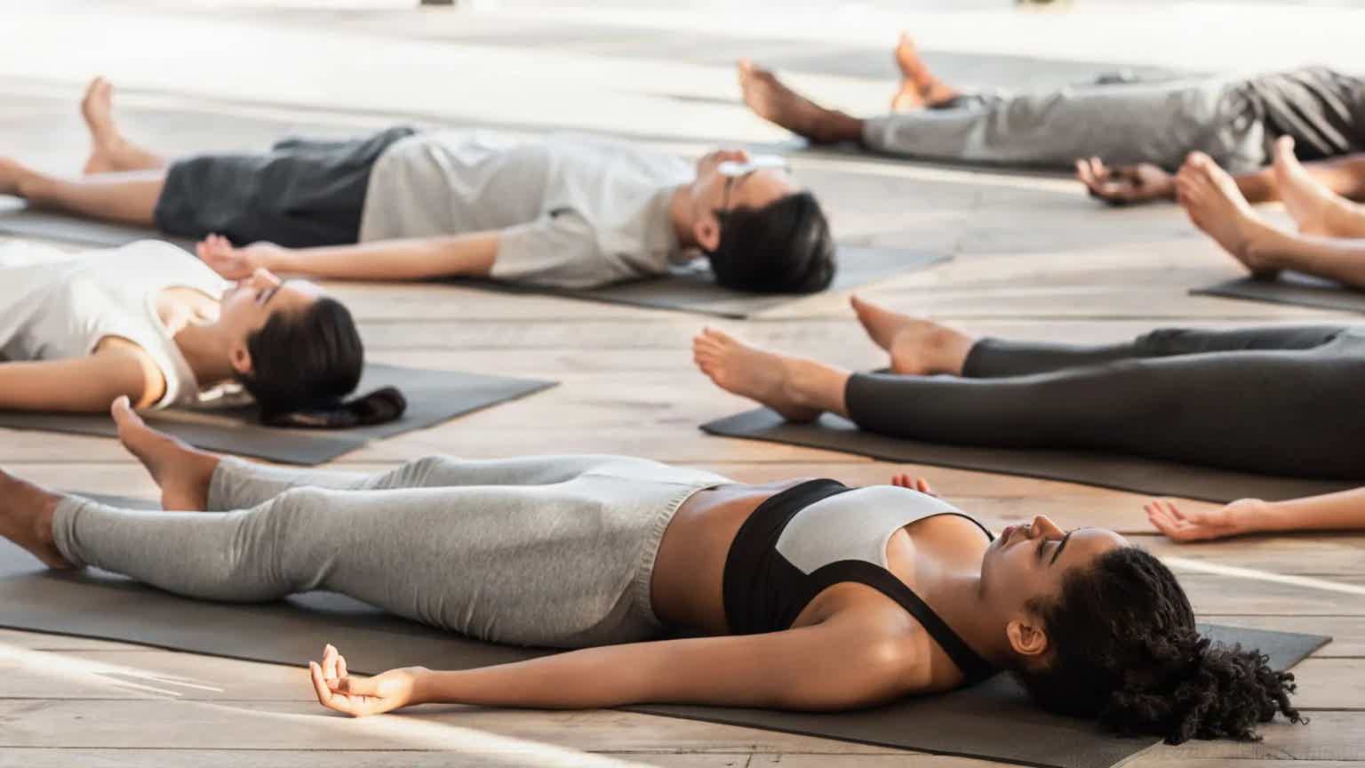 Group yoga class in a studio, participants lying in supine pose with arms extended on mats.