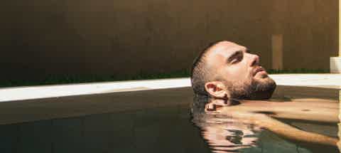 Bearded man relaxing in an outdoor pool, head and shoulders above water, eyes closed.