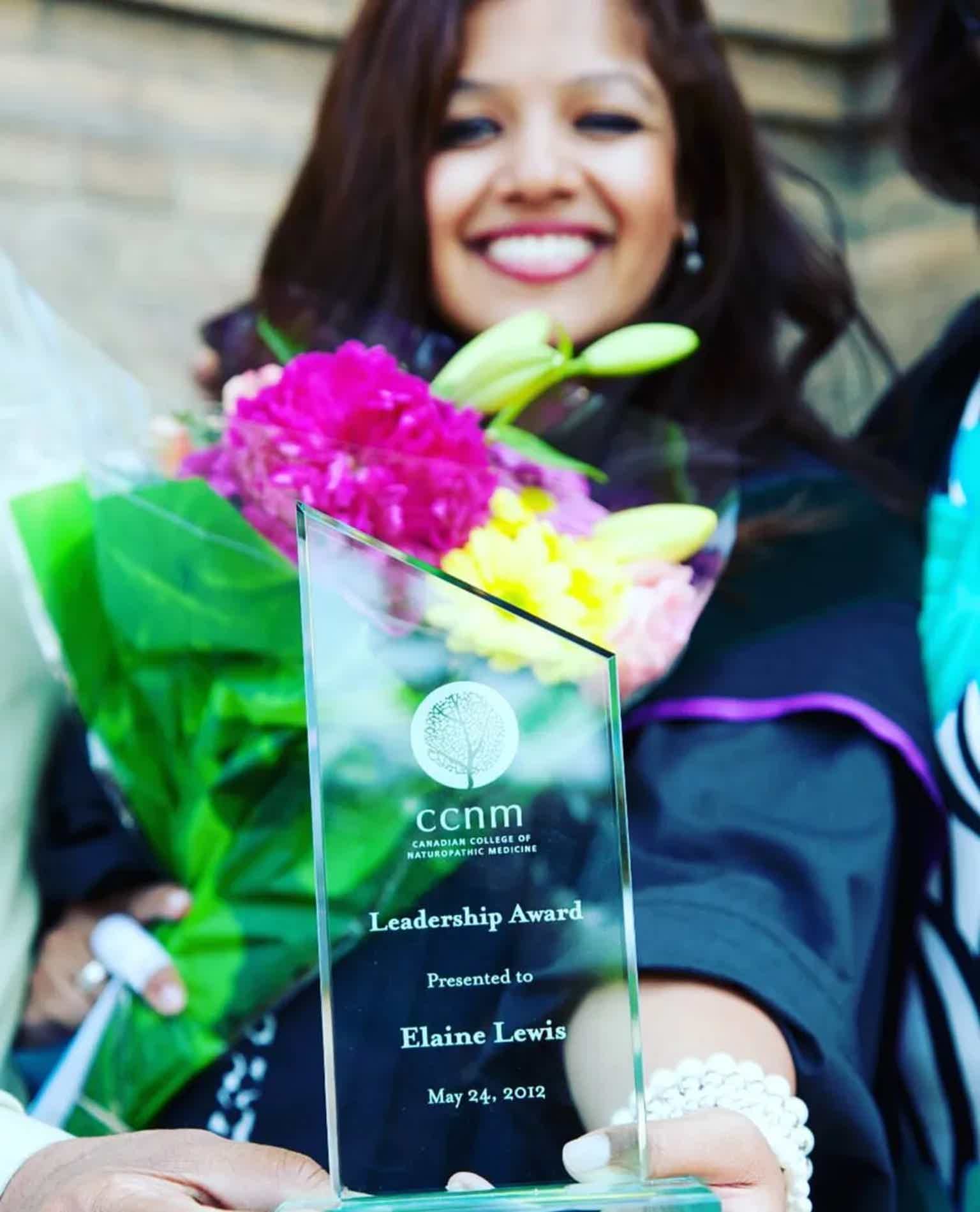 Crystal award with inscription, bouquet of flowers, smiling graduate in cap and gown.