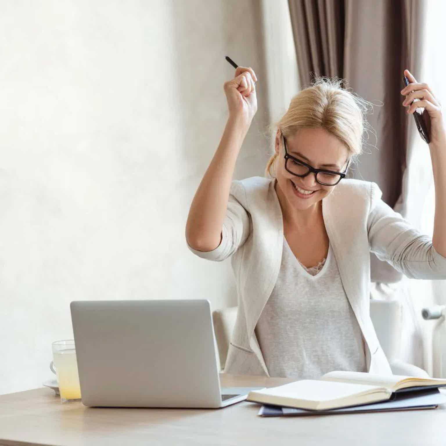 Smiling blonde woman in glasses raising arms as she works at a desk with laptop and open book near window curtains