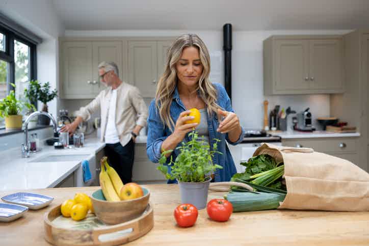 Woman in a bright kitchen pouring juice while holding a bunch of greens and preparing vegetables on the counter.