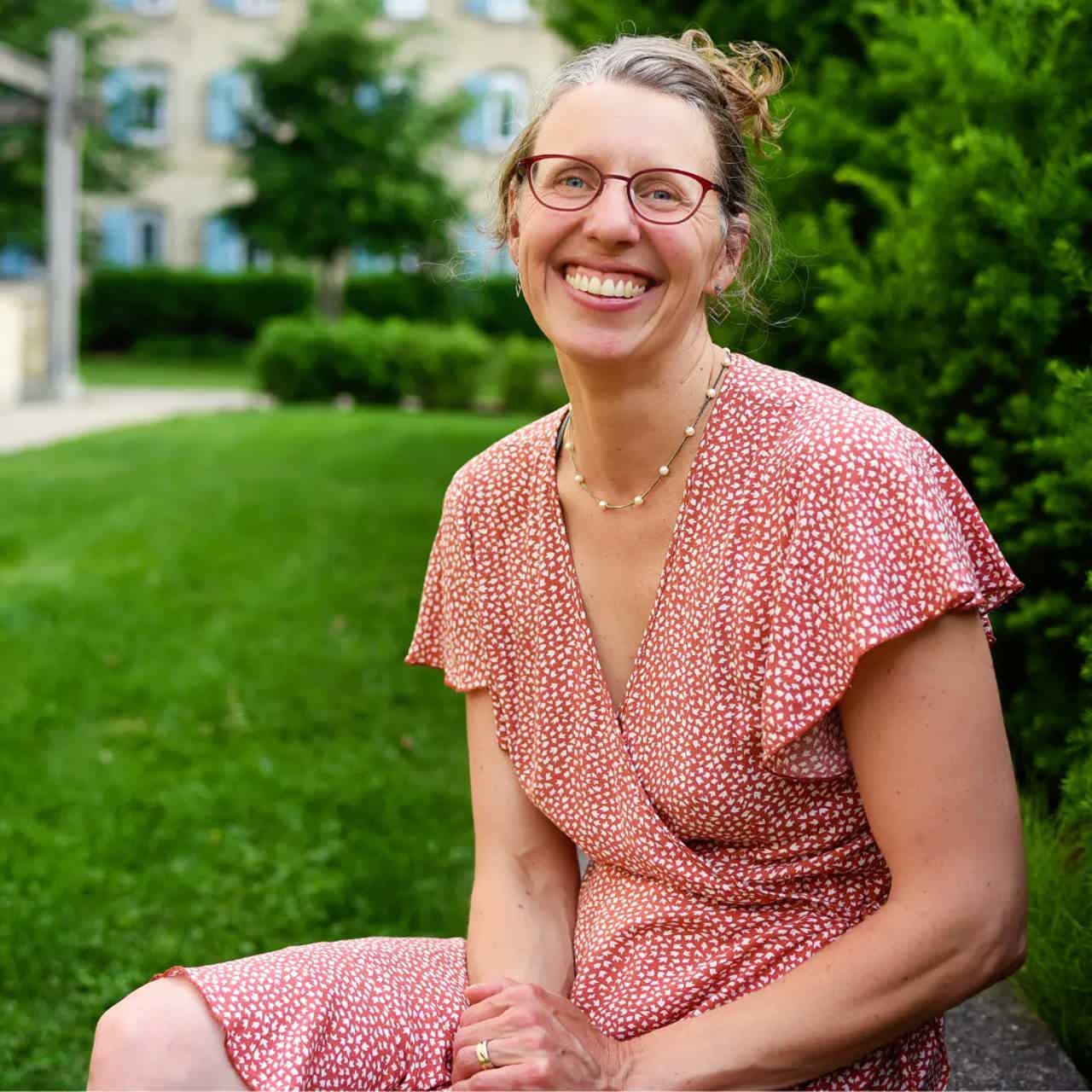 Smiling woman with glasses in a red patterned dress sitting on a bench in a green garden.