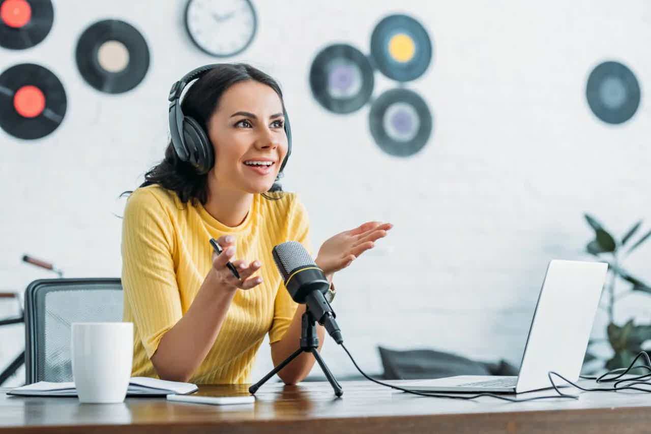 Smiling woman in yellow shirt wearing headphones and speaking into a studio microphone during a video call.