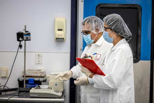 Two lab technicians in protective gear recording results beside laboratory testing equipment.