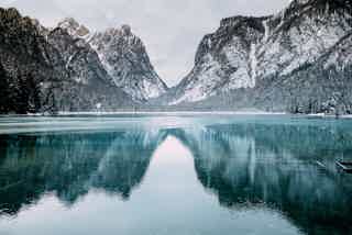 Snowy mountains rise over a calm, turquoise lake with a glassy reflection. Snow blankets peaks and shoreline.