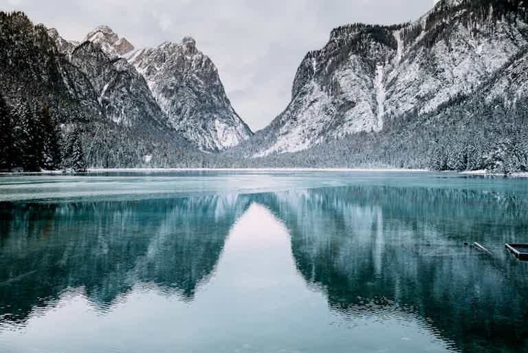 Snowy mountains rise over a calm, turquoise lake with a glassy reflection. Snow blankets peaks and shoreline.
