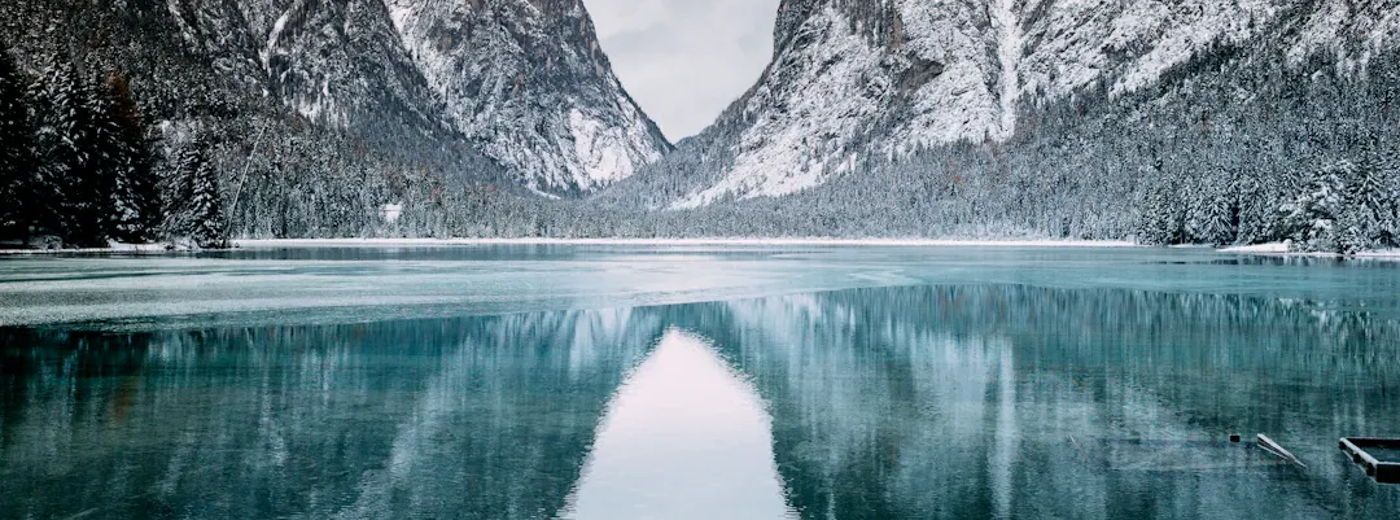 Snowy mountains rise over a calm, turquoise lake with a glassy reflection. Snow blankets peaks and shoreline.