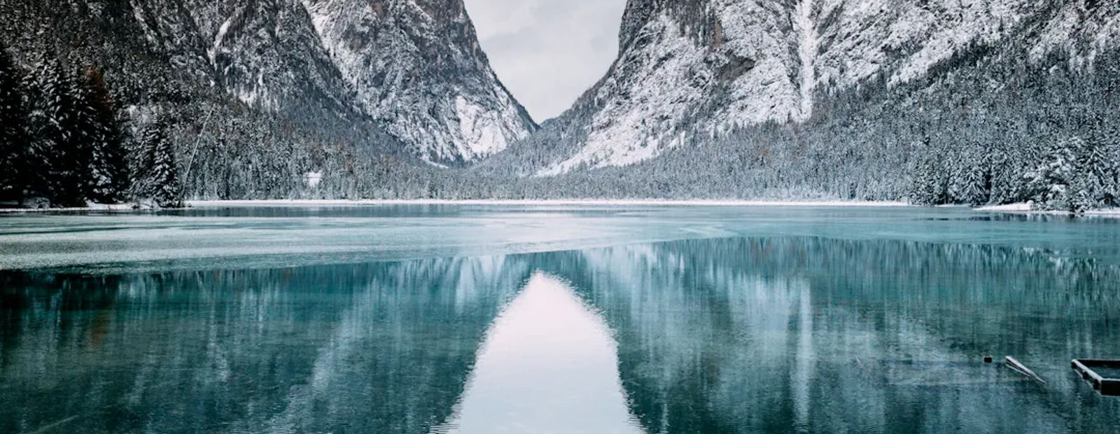 Snowy mountains rise over a calm, turquoise lake with a glassy reflection. Snow blankets peaks and shoreline.