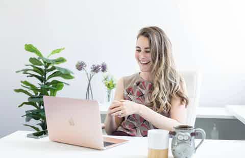 Smiling woman in floral dress working at a white table with laptop, coffee mug, and potted plant in bright room.