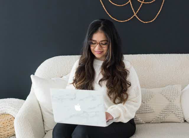 Smiling woman with long dark hair, white sweater, seated on pink chair, plant beside her in a bright room.