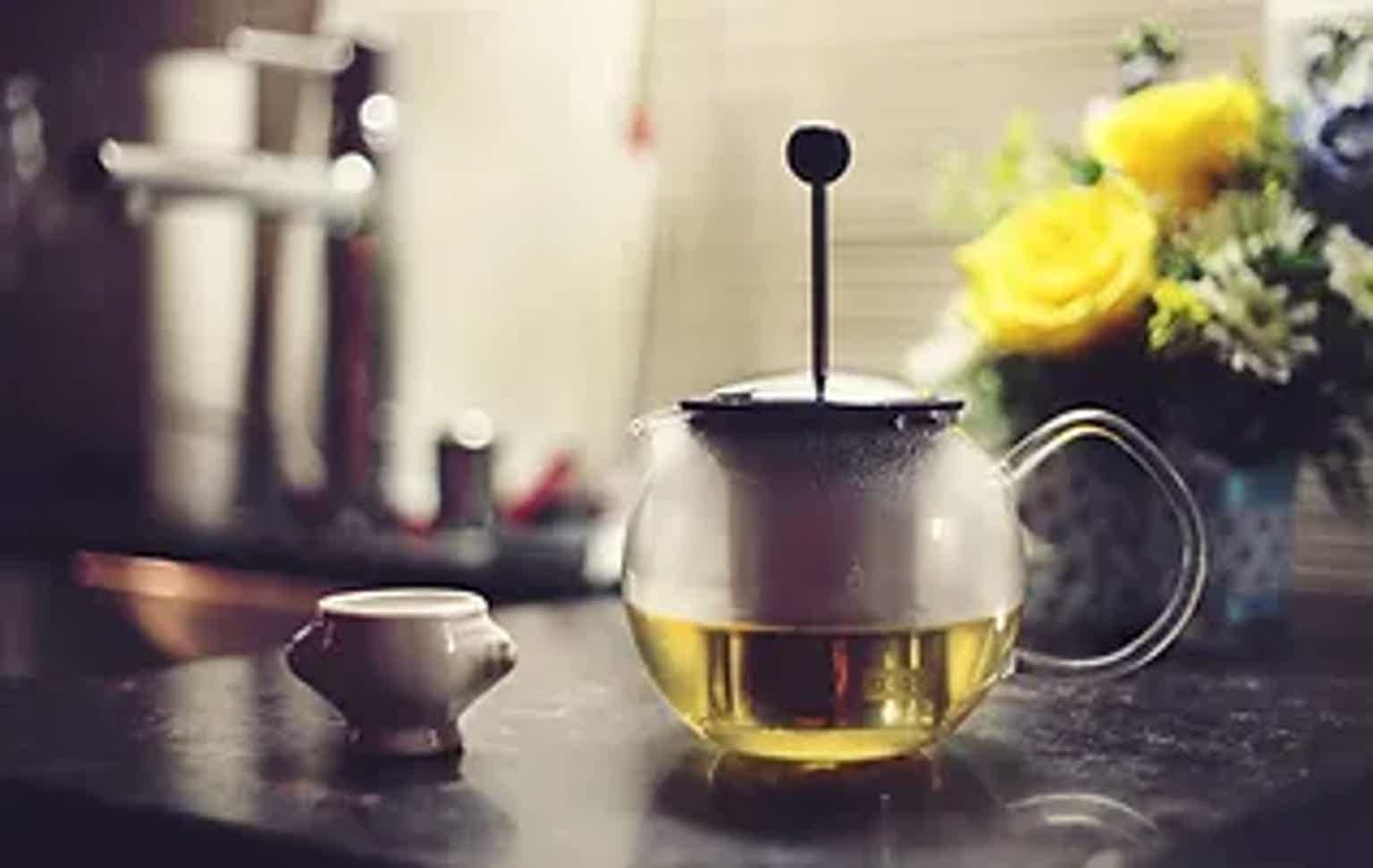 Glass teapot with yellow tea on a kitchen counter, small cup alongside, yellow flowers in background.