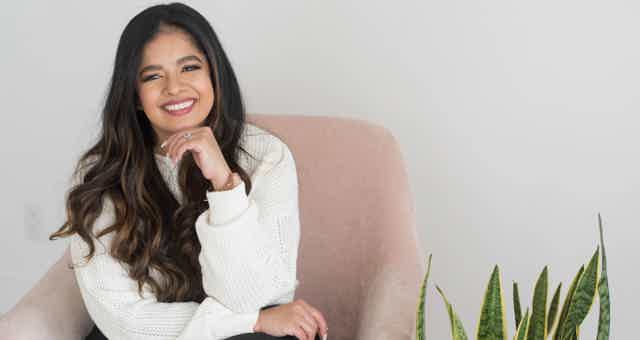 Smiling woman with long dark hair, white sweater, seated on pink chair, plant beside her in a bright room.