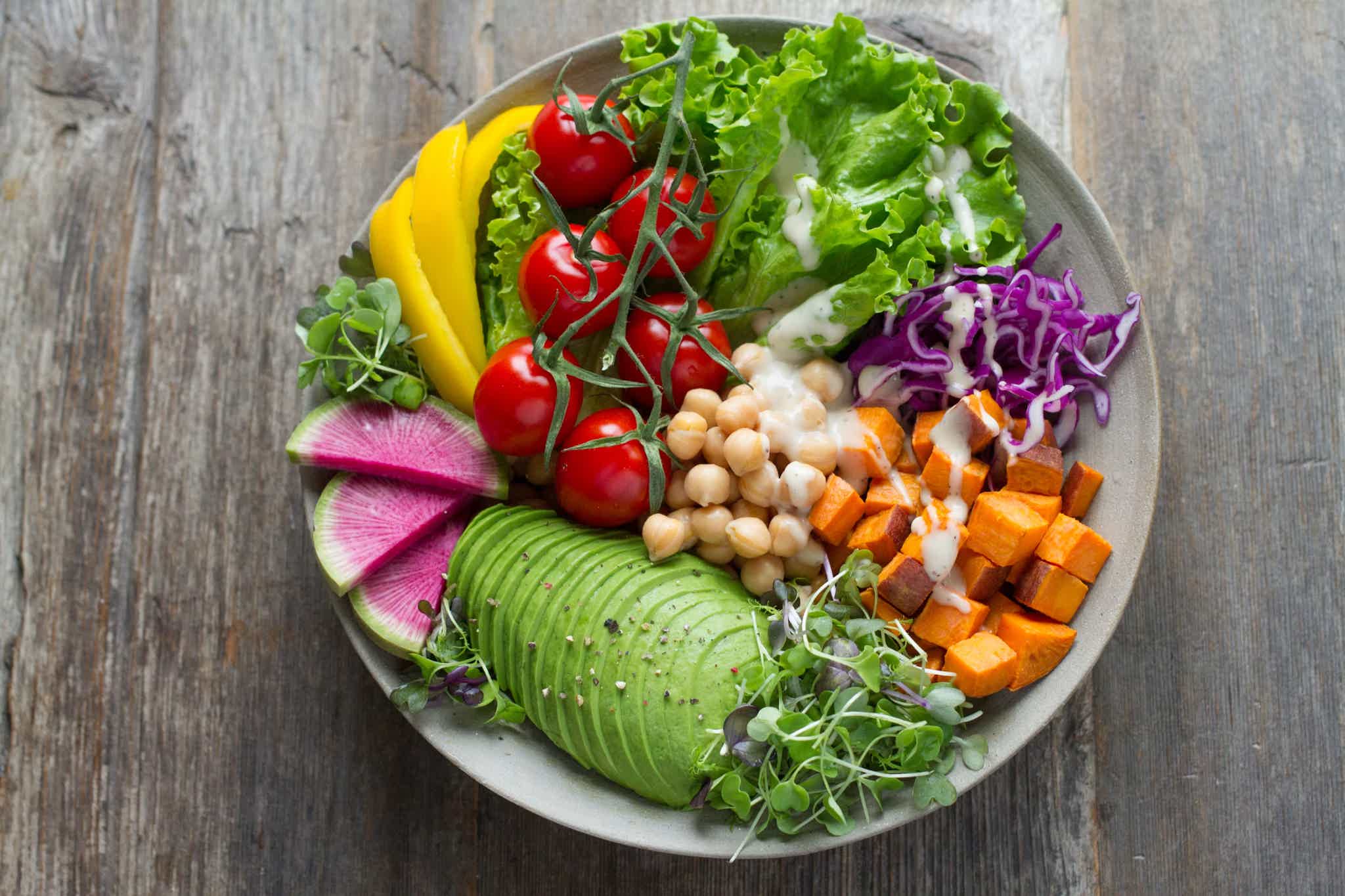 Colorful vegetable bowl with avocado, cherry tomatoes, chickpeas, sweet potato, radish, cabbage, and lettuce on wood.