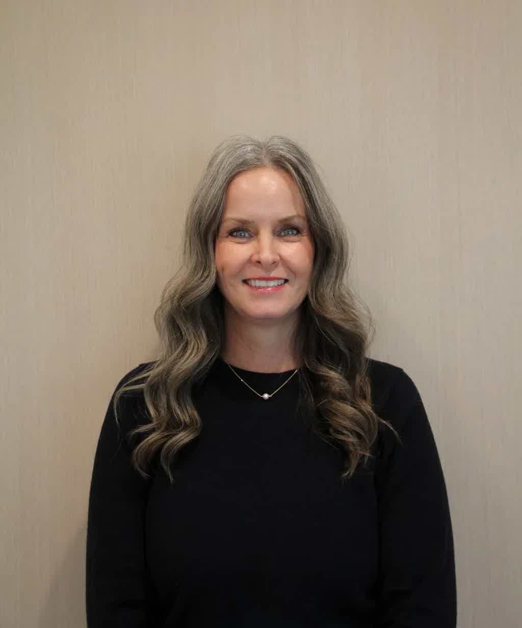 Smiling woman with wavy gray hair, black top, simple gold necklace, neutral beige background.