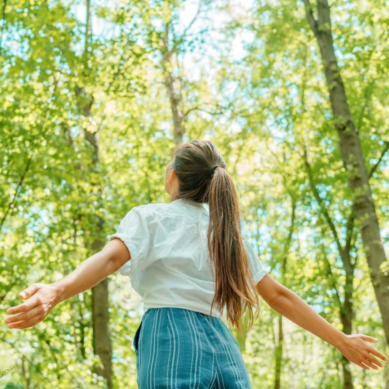 Woman in white blouse with long ponytail standing with arms open in a green forest.