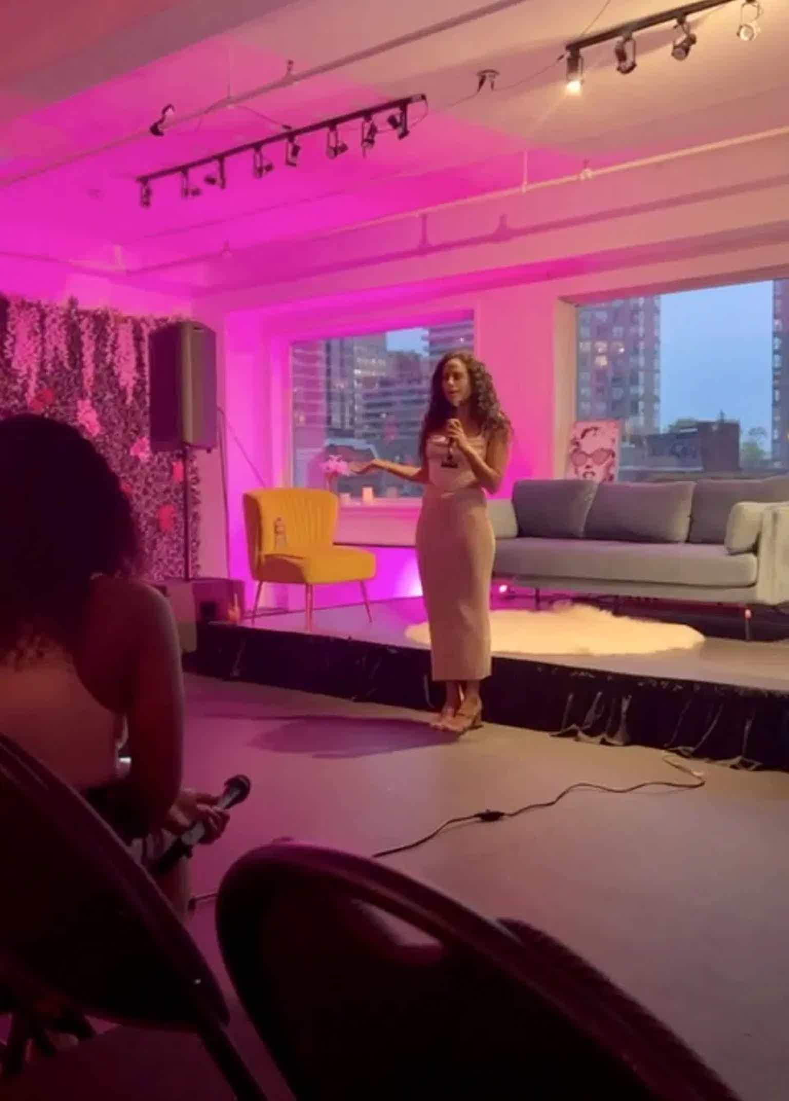 Woman presenting at an event with pink lighting in a modern indoor setting.