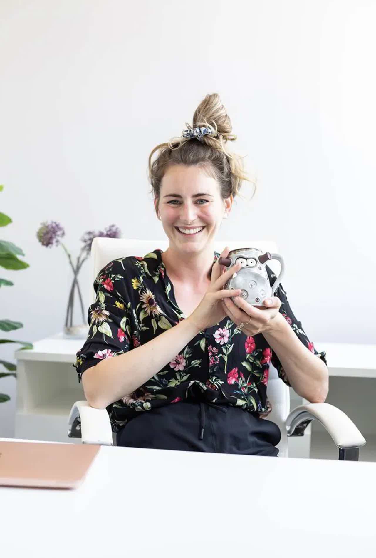Smiling woman in floral scrub top holding a patterned ear thermometer and taking a selfie indoors.