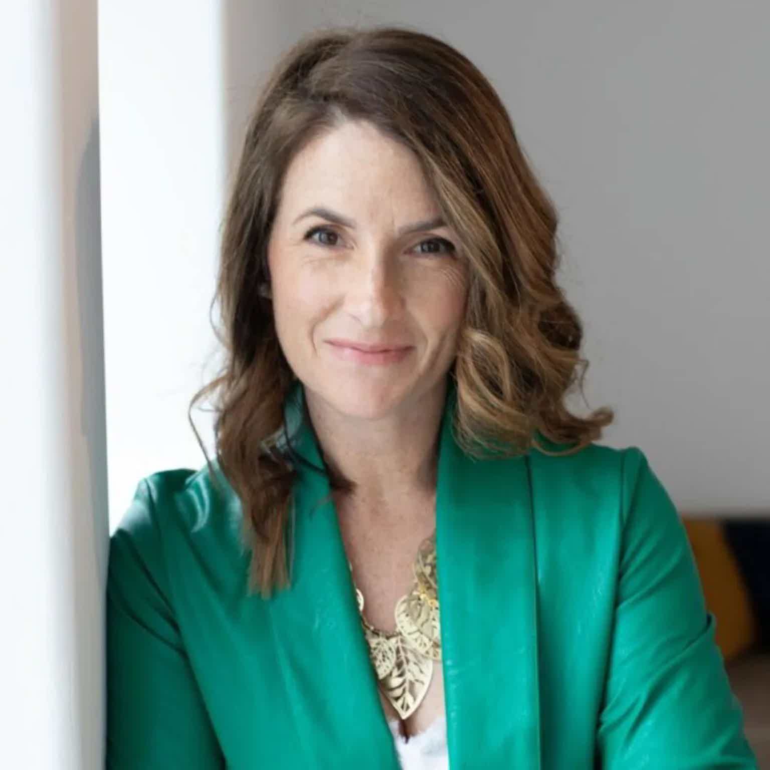 Smiling woman with shoulder-length wavy brown hair wearing a green blazer and gold necklace, facing the camera indoors.