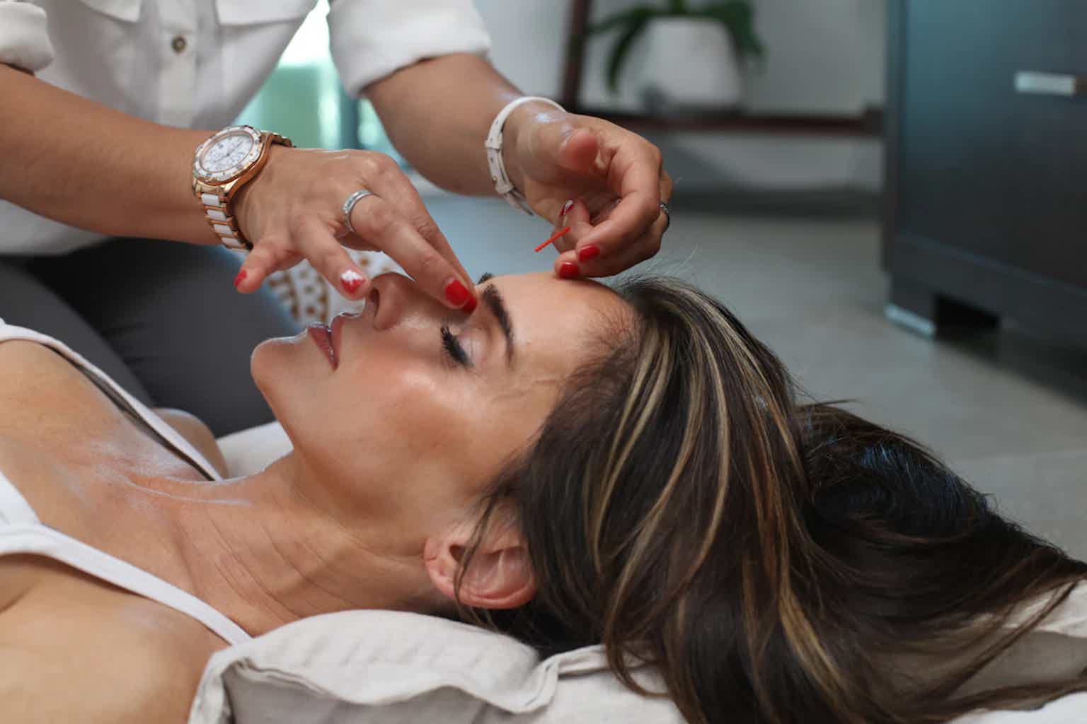 Esthetician applies facial treatment, placing small red and black tools on a client’s face in a clinic.