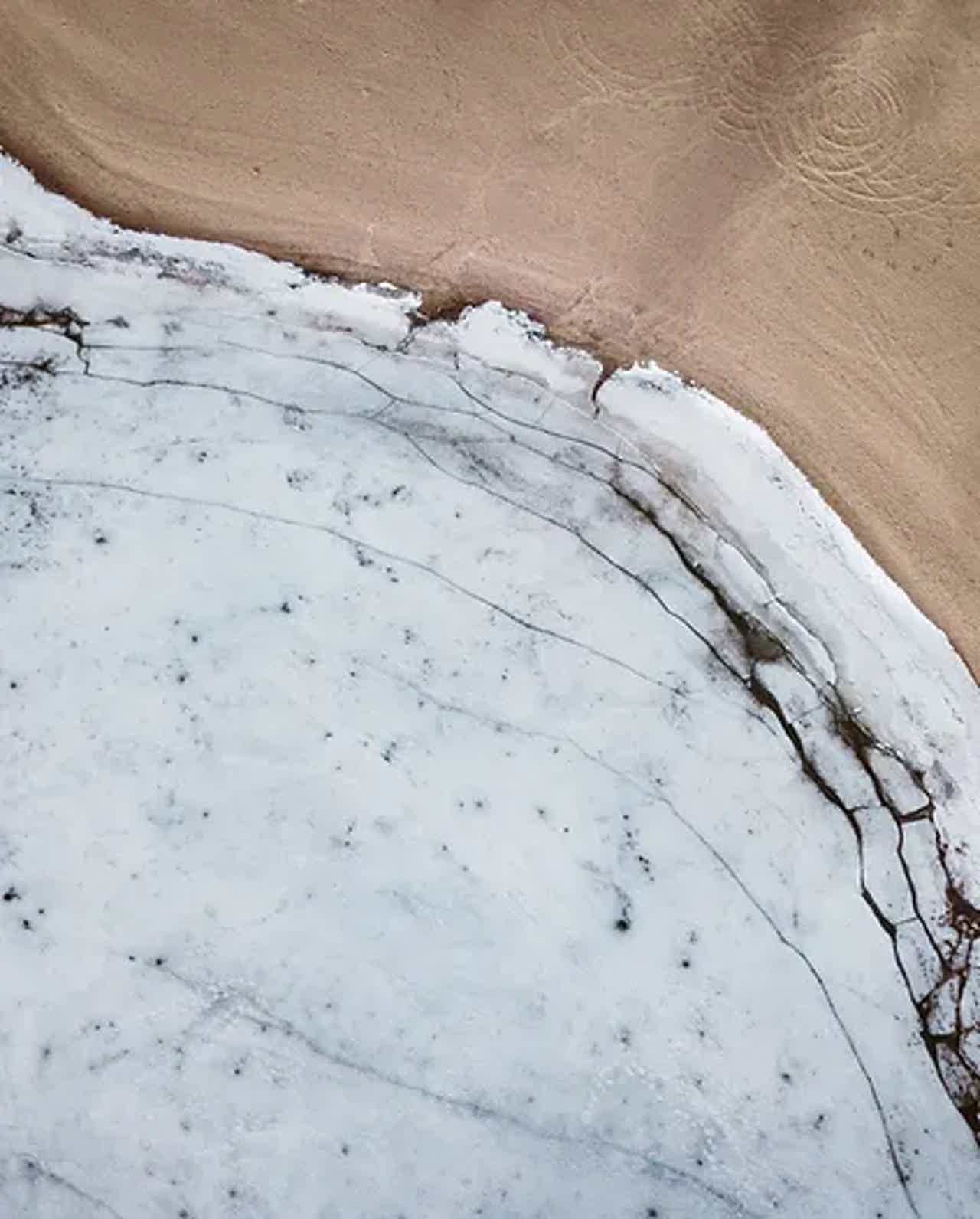 Aerial view of a snow-covered area with cracked ice edging towards sandy ground.