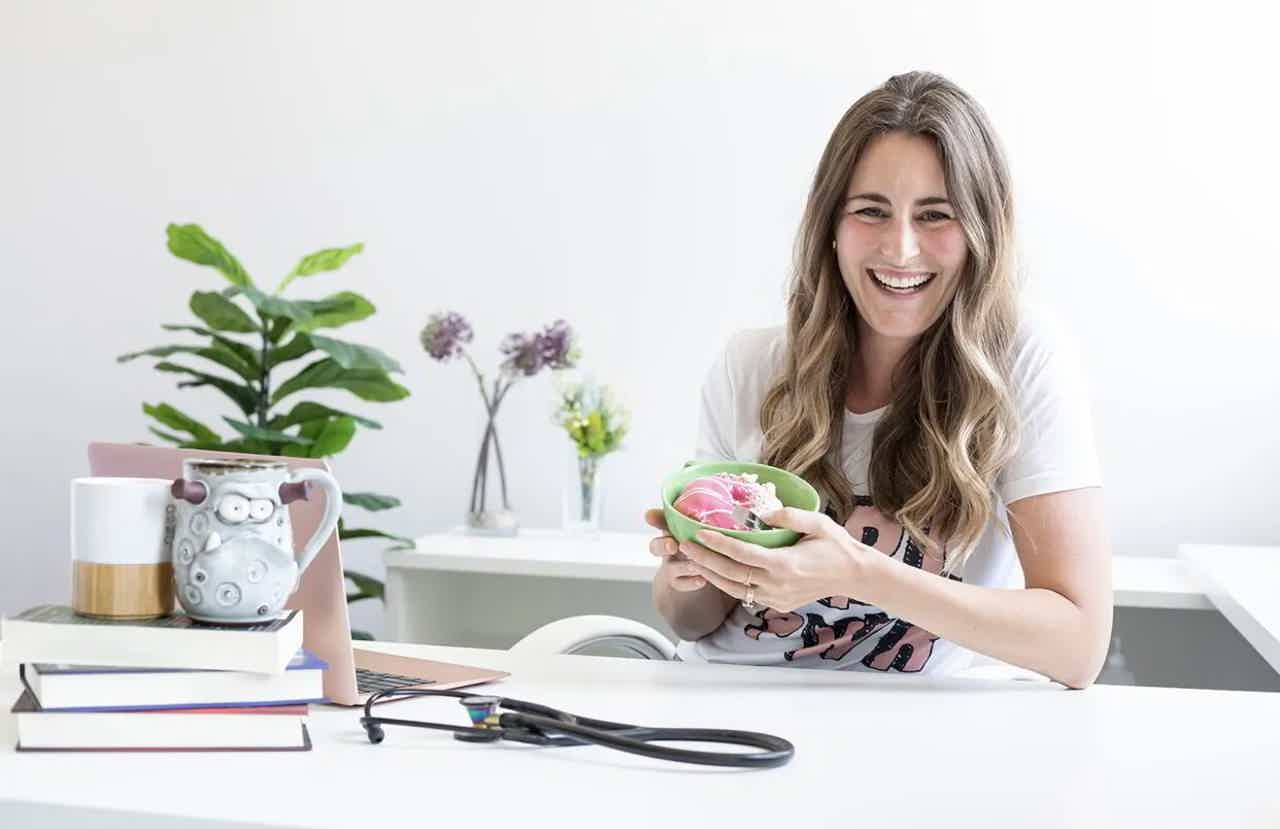 Smiling woman holding a small pink-and-green makeup brush holder at a white desk with flowers, glasses, and scissors