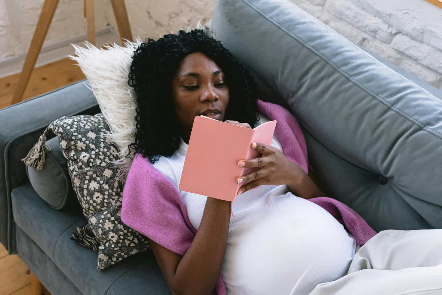 Woman reclining on teal couch, holding a pink book and reading; pillows and cozy blanket nearby.
