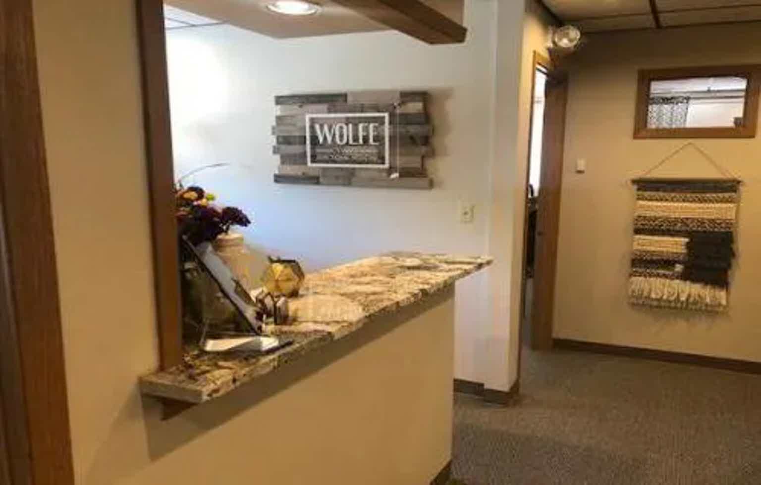 Wall fireplace mantel with “WOLFE” sign, flowers on shelf, framed textiles hanging in hallway.