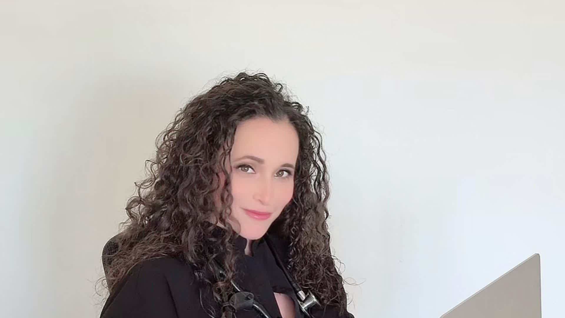 Person with long curly dark hair wearing a black top, standing behind a microphone against a white background.